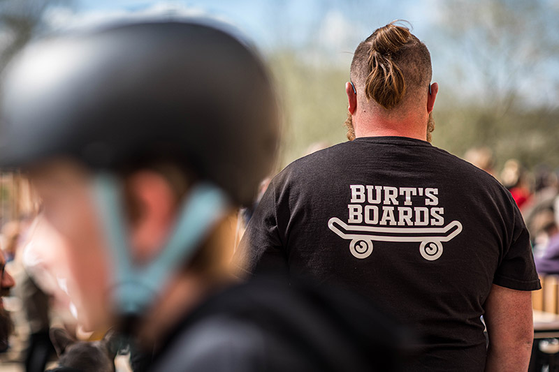 We see a Burt's Board's employee from the back wearing a Burt's Boards t-shirt. In the foreground is a blurry skateboarder with a helmet on
