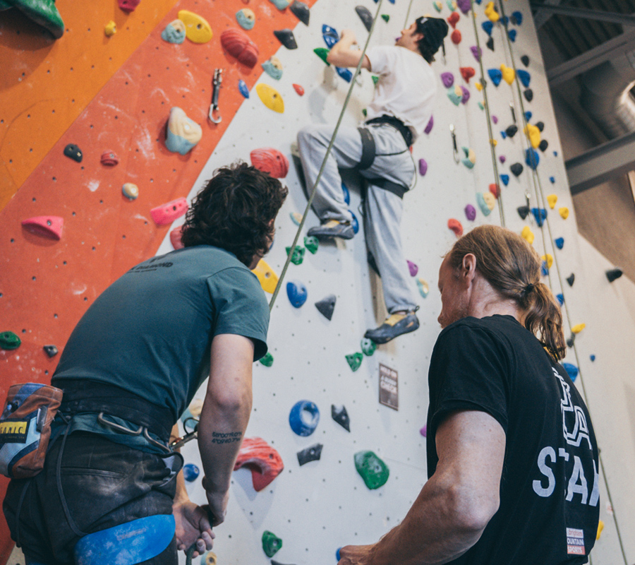 An instructor teaches someone in the foreground to belay while their friend is climbing in the background.