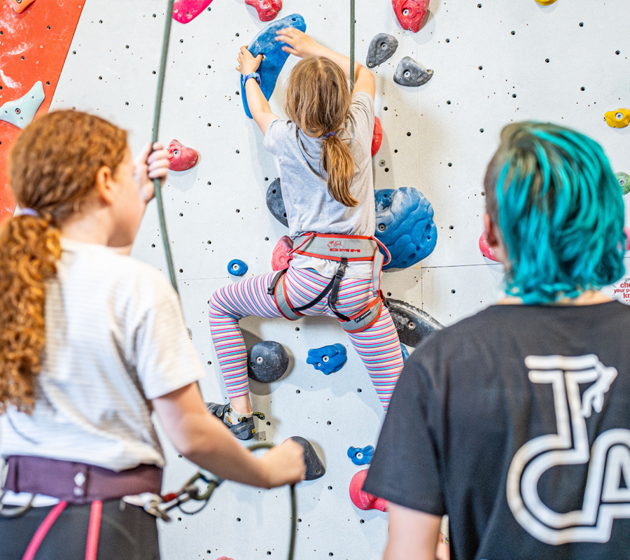 A young girls belays her friend who is climbing while an instructor sits with them and holds the ropes