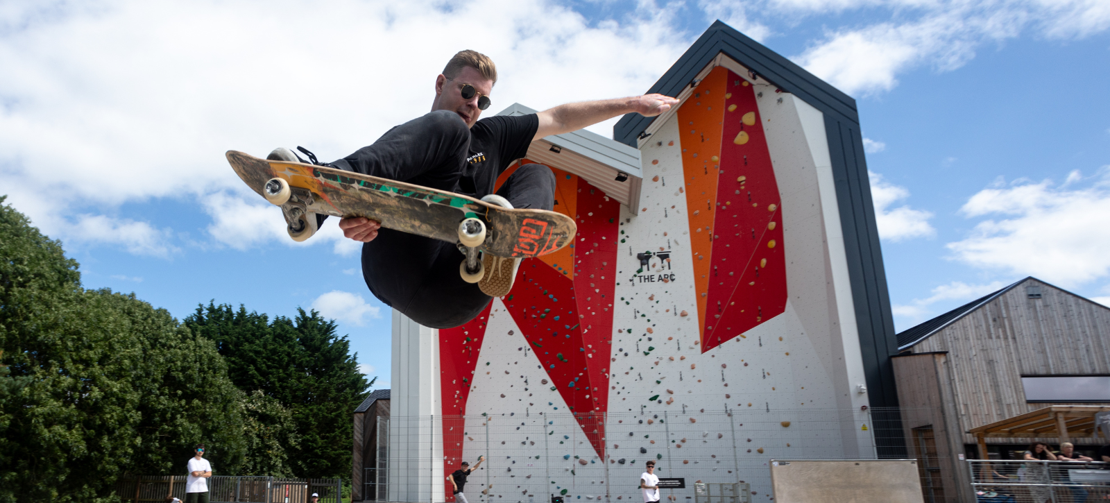 A skateboarder does a trick suspended off the ground in front of The Arc's outside climbing wall